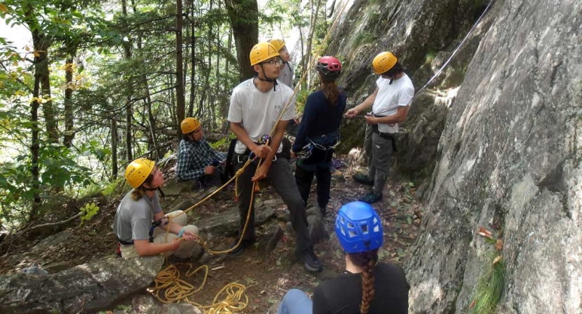 A group of people wearing helmets and other safety gear gather at the base of a rock wall, preparing to climb